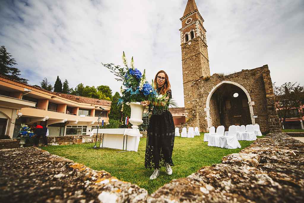 Poročna načrtovalka Petra Starbek nosi belo renesančno vazo z belo-modrim cvetjem v Bernardinu. Foto: Aleks in Irena Kus Wedding Photography