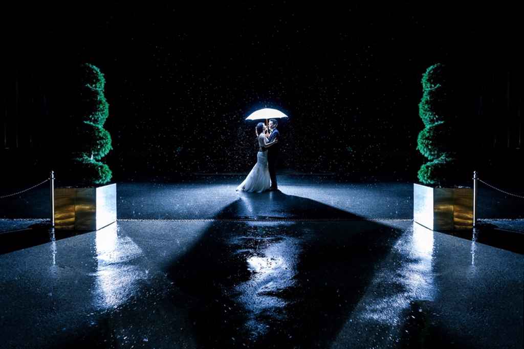 Nočna fotografija para, ki stoji zunaj tesno objet pod belim dežnikom pred kongresnim centrom Brdo. Foto: Ana Gregorič Photography