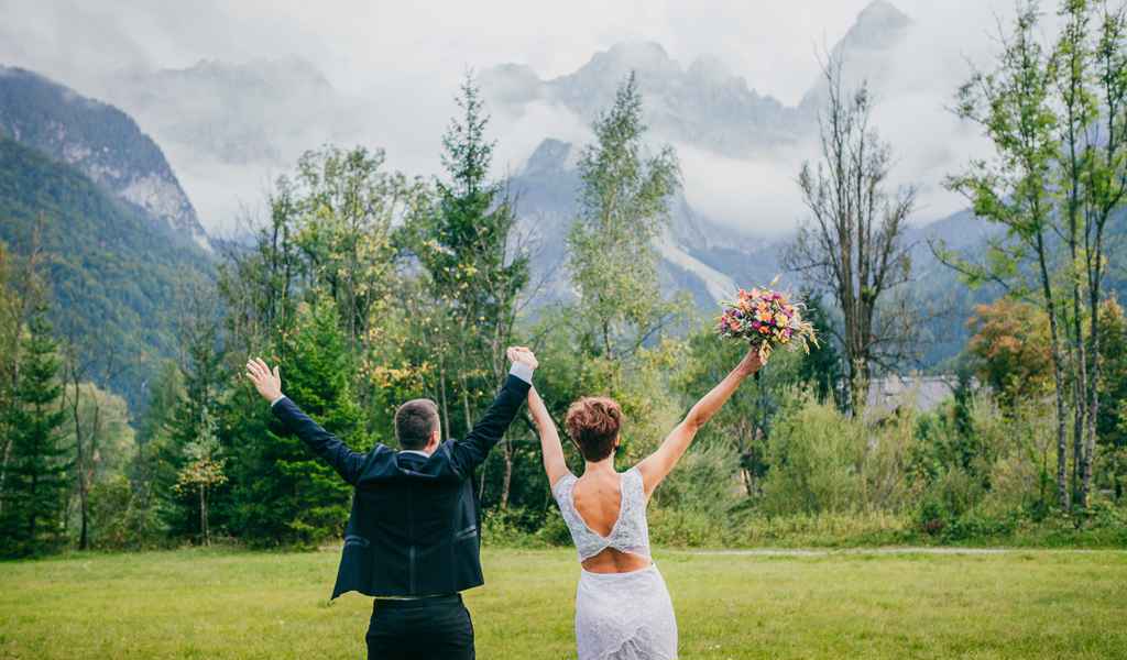 Nevesta s poročnim šopkom iz cvetličarne Emporija in ženin se sprehajata z dvignjenima rokama v gozdu Martuljek. Foto: Polona Bartol Photography