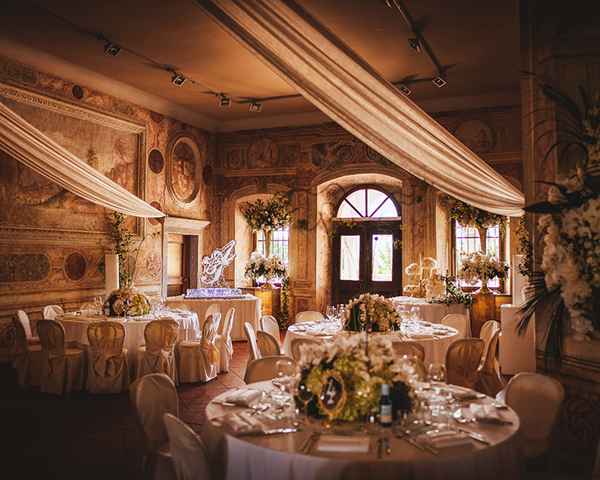 Luksuzna cvetlična poročna dekoracija v prekrasnem mogočnem dvorcu Zemono v Vipavi. Okrogle mize krasi cvetlična dekoracija v zeleno beli barvi ter številke miz na zlatih ogledalih. S stropa proti vratom se spuščajo trakovi belega blaga.