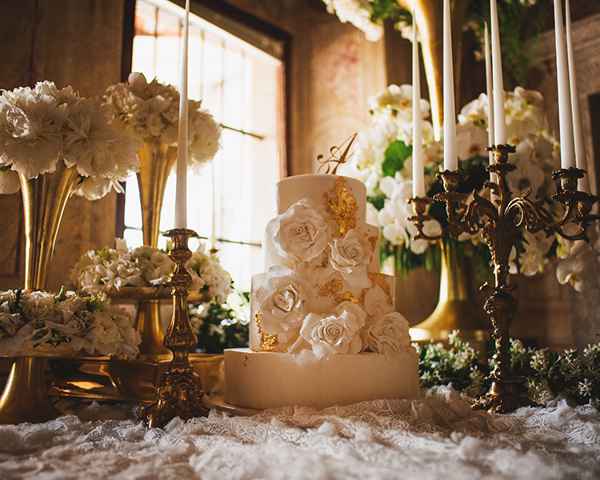 Prečudovita unikatna štirinadstropna bela poročna torta z zlatimi lističi in ročno izdelanimi cvetovi. Na vrhu je zlat monogram. Torta je postavljena v središče cvetlične dekoracije na sladkem omizju. 